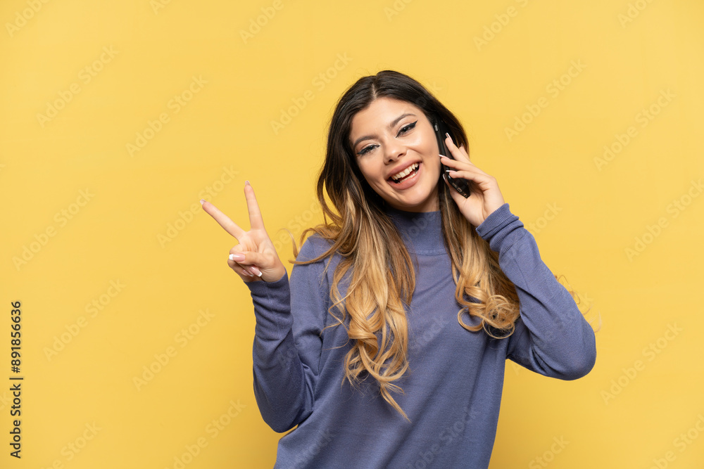 Young Russian girl using mobile phone isolated on yellow background smiling and showing victory sign Young Russian girl using mobile phone isolated on yellow background smiling and showing victory sign