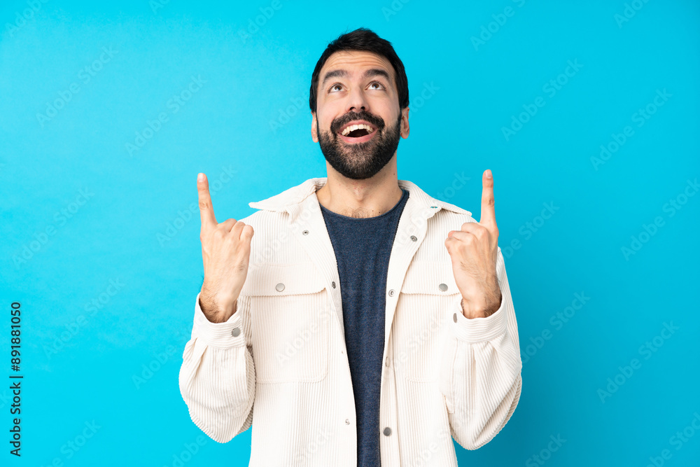 Young handsome man with white corduroy jacket over isolated blue background surprised and pointing up Young handsome man with white corduroy jacket over isolated blue background surprised and pointing up