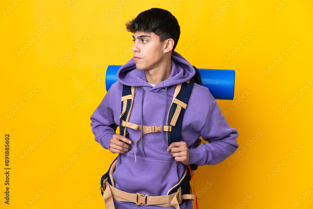 Young mountaineer man with a big backpack over isolated yellow background looking to the side Young mountaineer man with a big backpack over isolated yellow background looking to the side