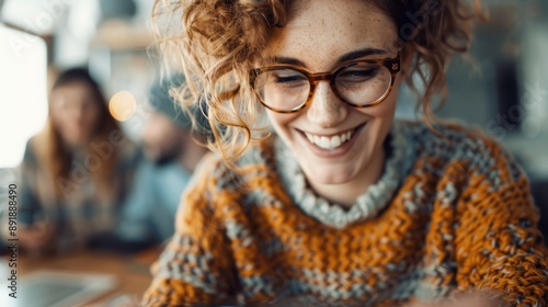 A woman with curly hair and glasses beams with joy as she focuses on her work or leisure activity, all while wearing a cozy, colorful knit sweater in a welcoming setting.