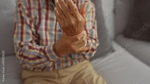 An elderly man in a plaid shirt, sitting on a couch, clutches his wrist in pain in the living room of his home. photo