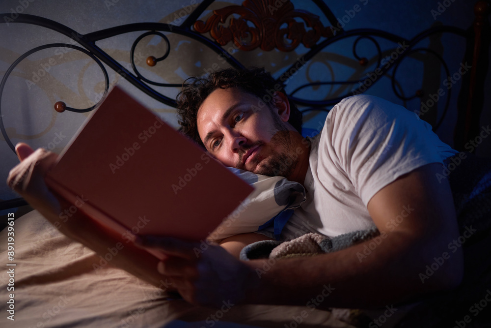 Fototapeta premium A man is peacefully reading a book in bed in a dimly lit bedroom, covered with cozy blankets