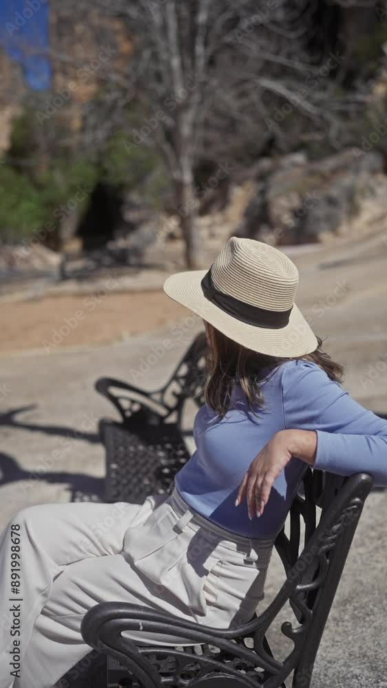 A smiling woman sits on a bench wearing a straw hat and casual clothes in a sunlit park. A smiling woman sits on a bench wearing a straw hat and casual clothes in a sunlit park.