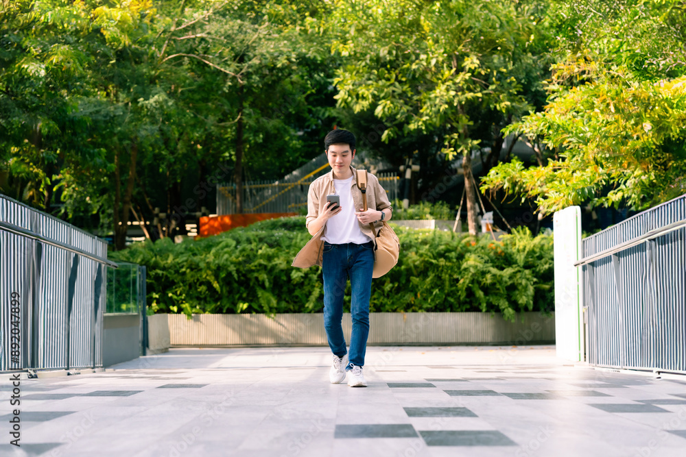 Fototapeta premium Portrait of handsome Asian student using smartphone. A young man walking outdoor happy smiling with holding mobile phone