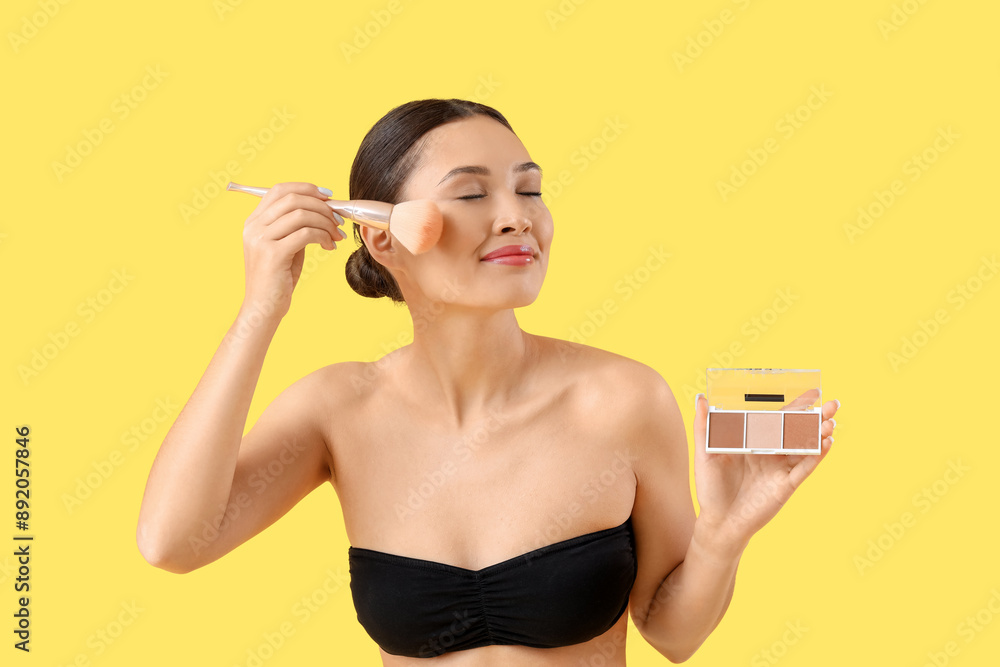 Fototapeta premium Young asian woman applying face powder with makeup brush and contour palette on yellow background, her eyes are closed in concentration, and she wears a strapless top