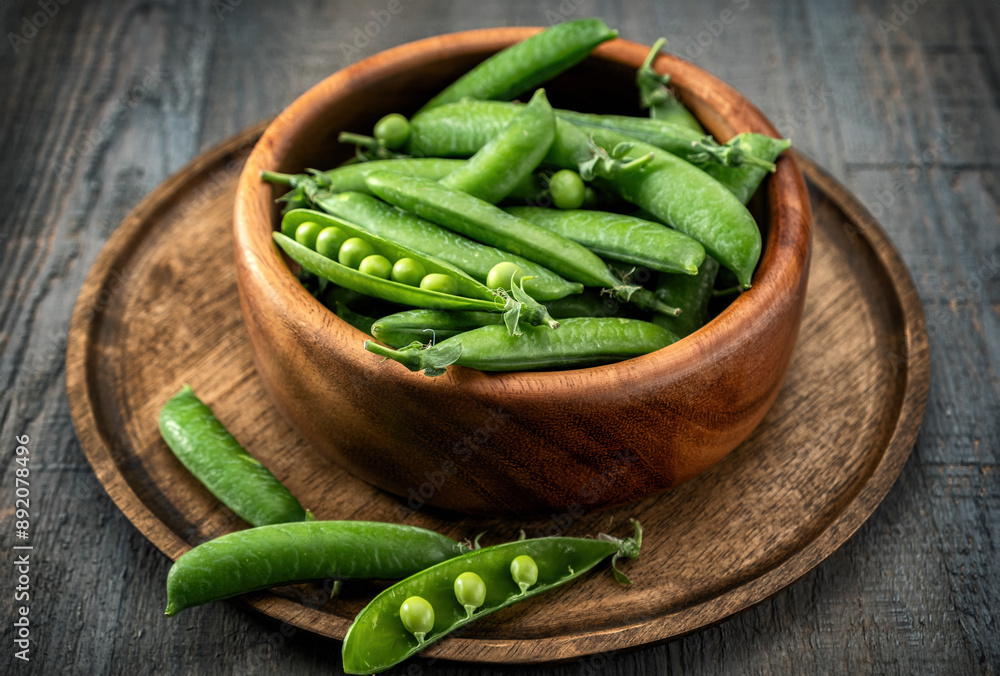 General plan of green young peas in pods in a wooden bowl on a wooden tray, on a gray textured wooden table. Low key. General plan of green young peas in pods in a wooden bowl on a wooden tray, on a gray textured wooden table. Low key.