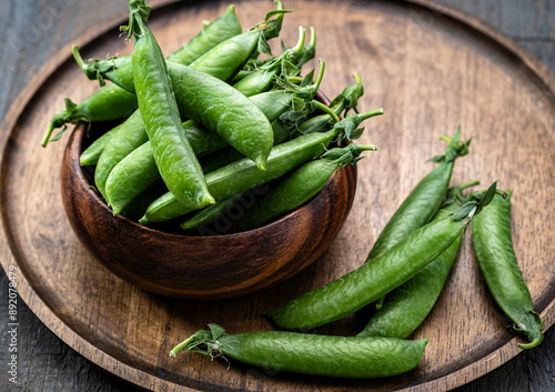 Green pea pods are piled in a wooden bowl on a round wooden tray. Rustic, low key.
