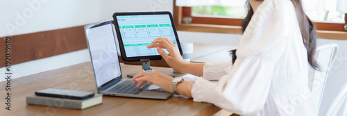 Young asian businesswoman checking market data of business on tablet and typing information on laptop while analysis strategy about investment and planning of new startup project to working in cafe