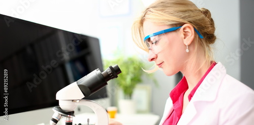 Female millennial chemist in protective gogles looking at microscope aganist chemistry lab background. Pharmaceuticals research panacea concept. photo