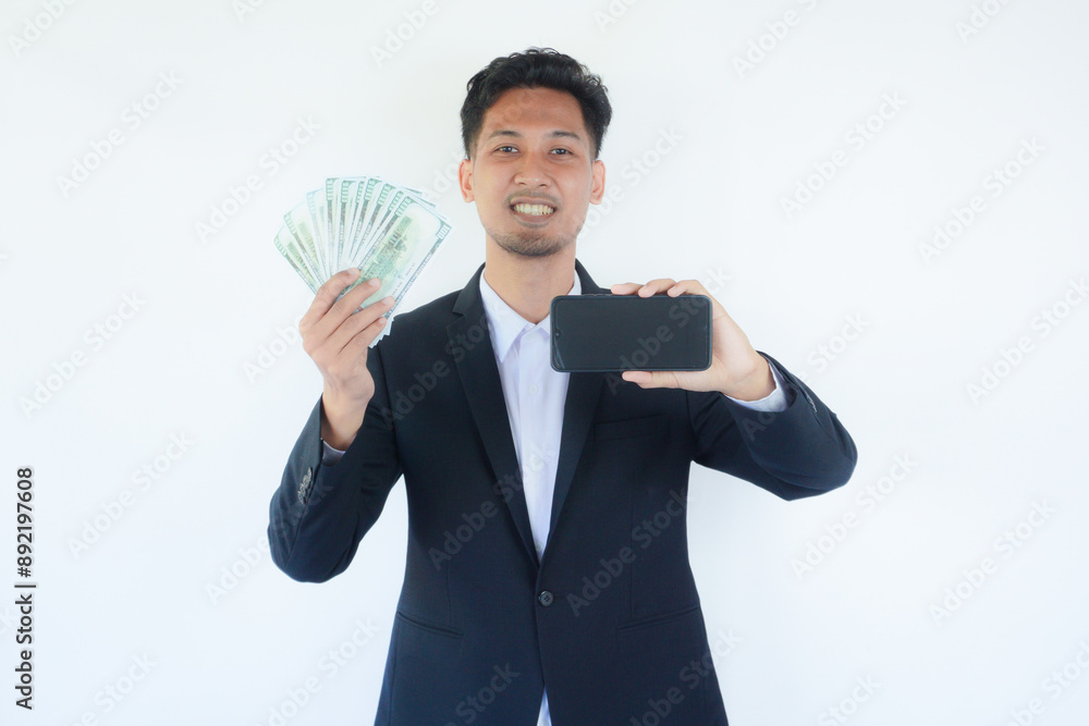 Portrait of an Indonesian Asian man, wearing a black suit, showing off the blank screen of his smartphone while holding money, isolated against a white background. Portrait of an Indonesian Asian man, wearing a black suit, showing off the blank screen of his smartphone while holding money, isolated against a white background.