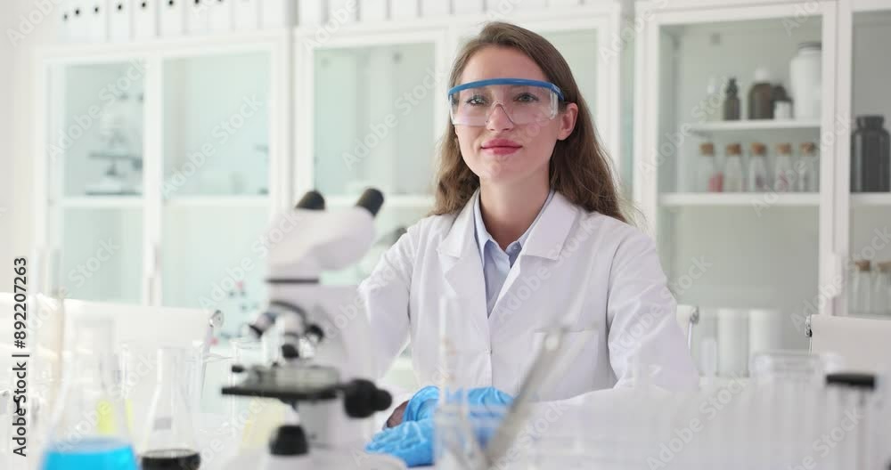 Smiling female lab technician sits at table with microscope. Medical worker at desk prepared to conduct experiments with cheerful expression Smiling female lab technician sits at table with microscope. Medical worker at desk prepared to conduct experiments with cheerful expression