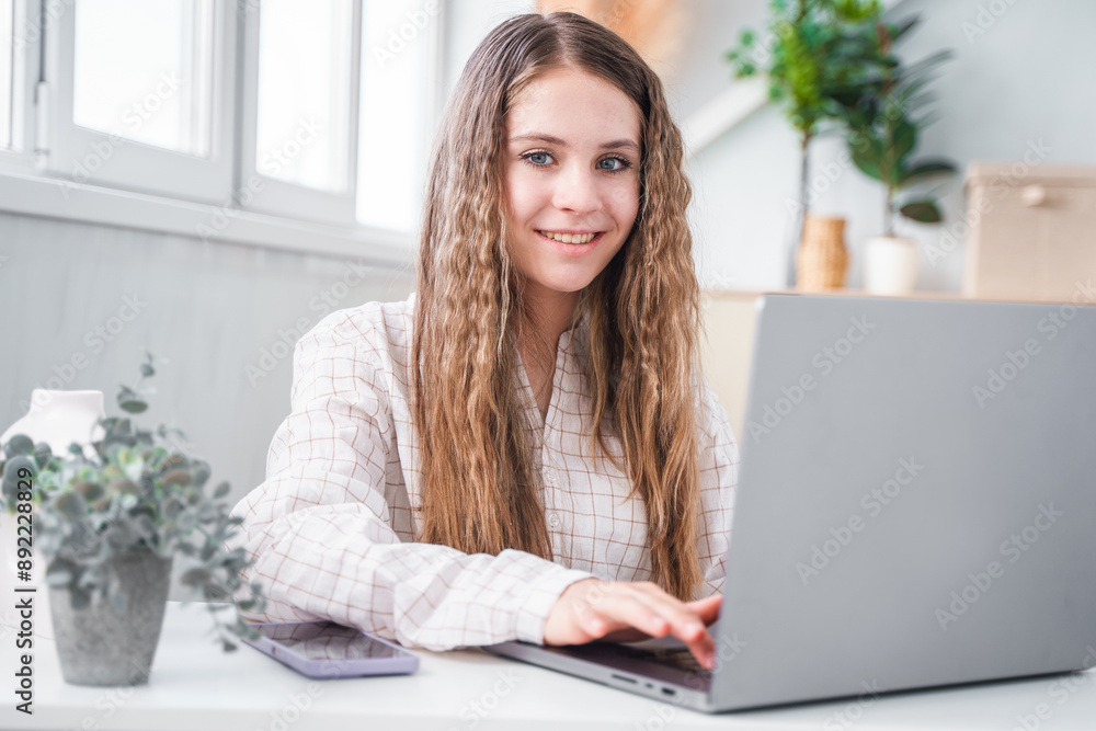 A young teenage student girl looking a laptop screen, online courses, studying remotely from home, sitting at a desk with a phone and laptop in a modern bright interior with plants. A young teenage student girl looking a laptop screen, online courses, studying remotely from home, sitting at a desk with a phone and laptop in a modern bright interior with plants.