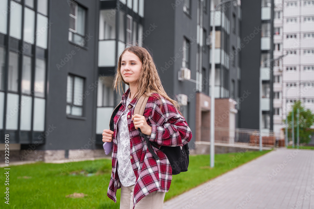 A young teenage girl with a backpack in a plaid shirt walks along an empty street between high-rise buildings. Teenage high school student girl with backpack on city street A young teenage girl with a backpack in a plaid shirt walks along an empty street between high-rise buildings. Teenage high school student girl with backpack on city street