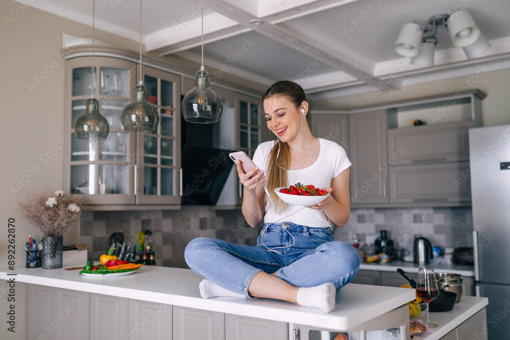 Photo of nice cheerful woman using cellphone and earphones while having breakfast in white kitchen Photo of nice cheerful woman using cellphone and earphones while having breakfast in white kitchen
