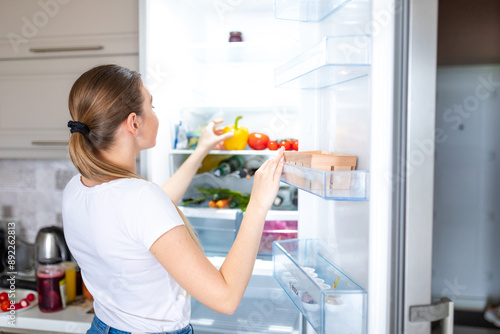 Konstfotografi Portrait of female standing near open fridge full of healthy food, vegetables and fruits
