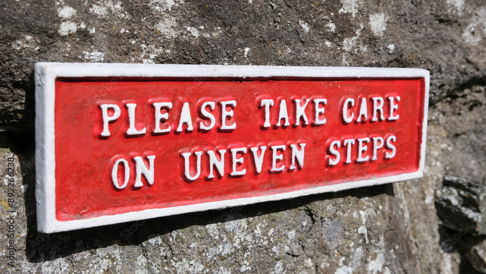 Red sign warning people to please take care on uneven steps against rustic stone wall Red sign warning people to please take care on uneven steps against rustic stone wall