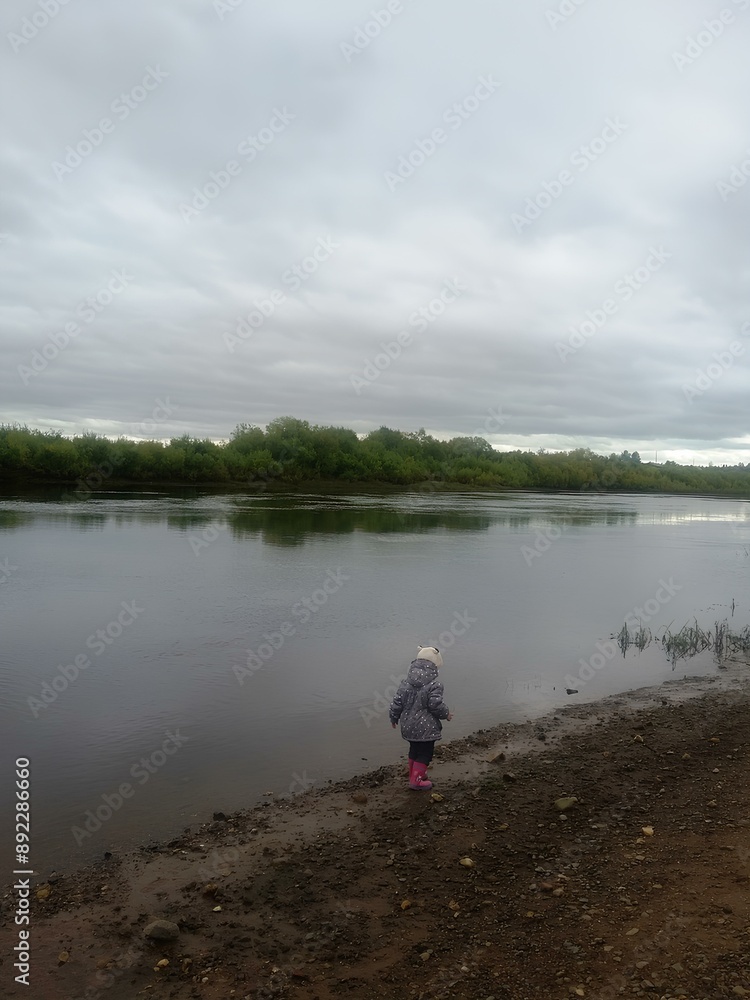a small child standing on the edge of a body of water, possibly a lake or river. The child is wearing a gray coat and a hat, and appears to be looking at the water a small child standing on the edge of a body of water, possibly a lake or river. The child is wearing a gray coat and a hat, and appears to be looking at the water