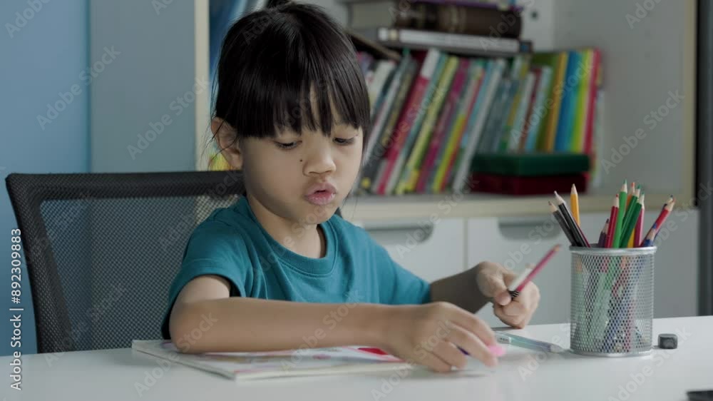 A cute Asian elementary school girl is sit and choosing her favorite colored pencils to draw and color in her favorite art book. She is happy spend her free time on the weekend. bookshelf background A cute Asian elementary school girl is sit and choosing her favorite colored pencils to draw and color in her favorite art book. She is happy spend her free time on the weekend. bookshelf background