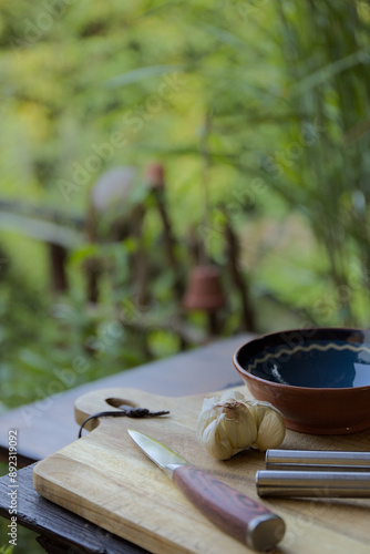Garlic cloves on a wooden board with a knife and a ceramic bowl in the garden with bokeh background