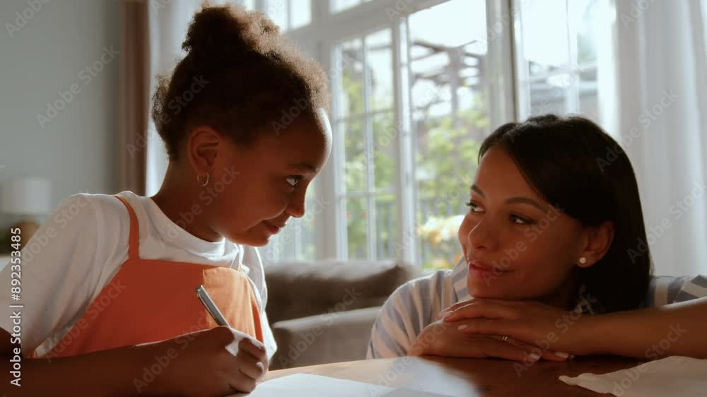African American schoolgirl is sitting at table with lamp, notebooks and pencils, writing and doing homework. Black skinned mother comes up to help and support her daughter African American schoolgirl is sitting at table with lamp, notebooks and pencils, writing and doing homework. Black skinned mother comes up to help and support her daughter