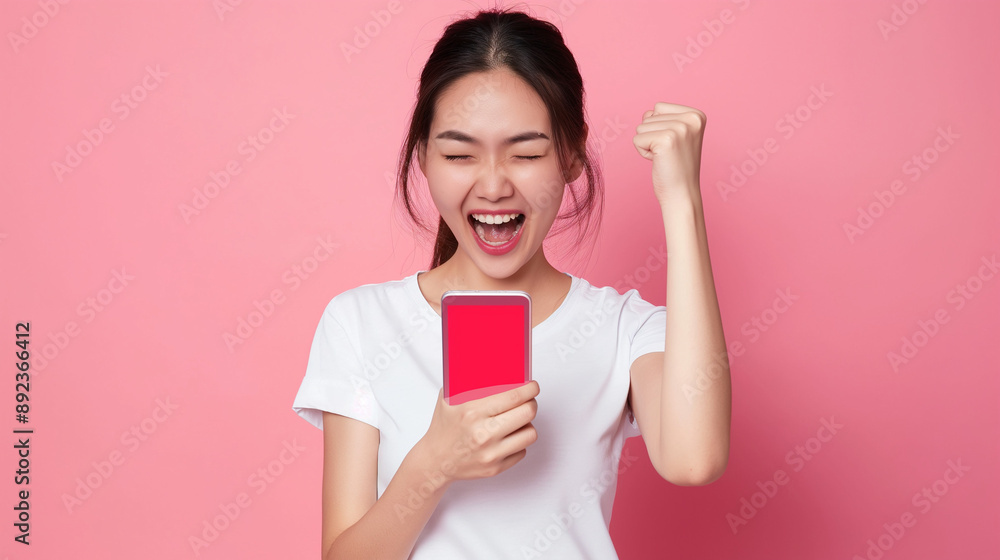 Young Asian woman in white t-shirt, excitedly celebrating with a smartphone against a pink background. Young Asian woman in white t-shirt, excitedly celebrating with a smartphone against a pink background.