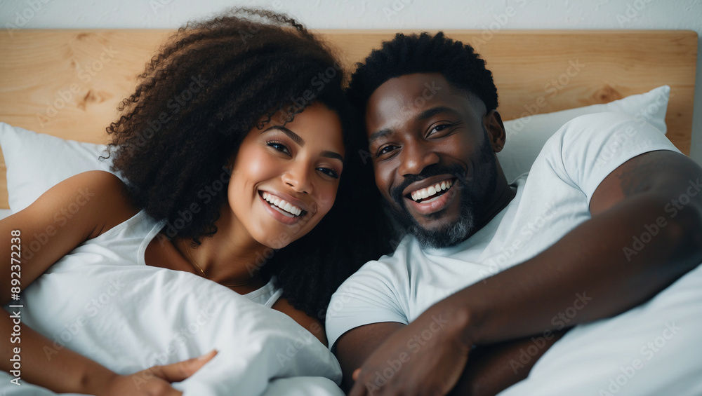 Smiling Black couple lies in bed, wrapped in white sheets and pillows. They are both looking directly at camera, with happy expressions on faces Smiling Black couple lies in bed, wrapped in white sheets and pillows. They are both looking directly at camera, with happy expressions on faces