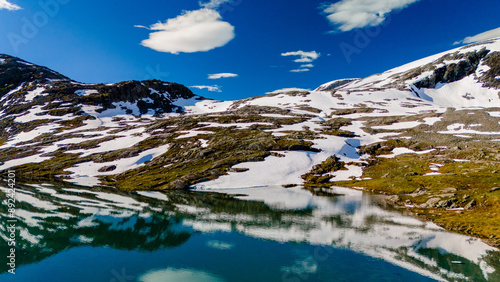 A picturesque mountain lake in Norway reflects the blue sky and white clouds above, surrounded by snow-capped peaks. Strynefjellsvegen, Geiranger, Norway photo