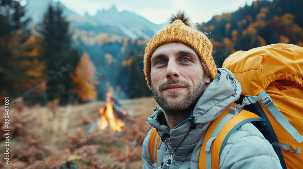 A man, wearing a yellow beanie and gray jacket, sits by a campfire among autumn trees in a mountainous region, with a yellow backpack beside him. A man, wearing a yellow beanie and gray jacket, sits by a campfire among autumn trees in a mountainous region, with a yellow backpack beside him.