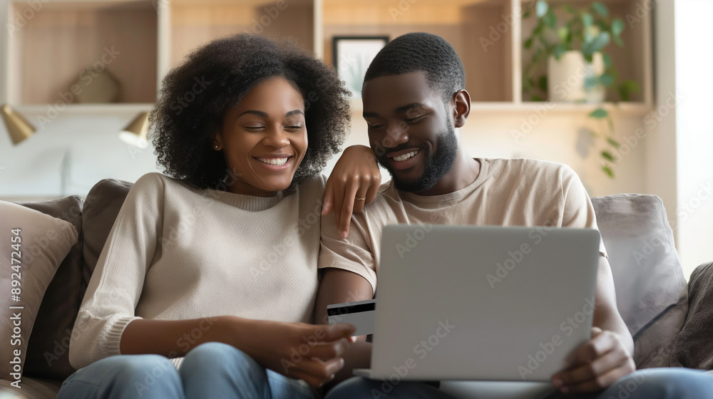 Cheerful young married couple, African American man and woman, sitting together on a sofa couch with a credit card and laptop, completing an e-commerce purchase, paying and buying Cheerful young married couple, African American man and woman, sitting together on a sofa couch with a credit card and laptop, completing an e-commerce purchase, paying and buying