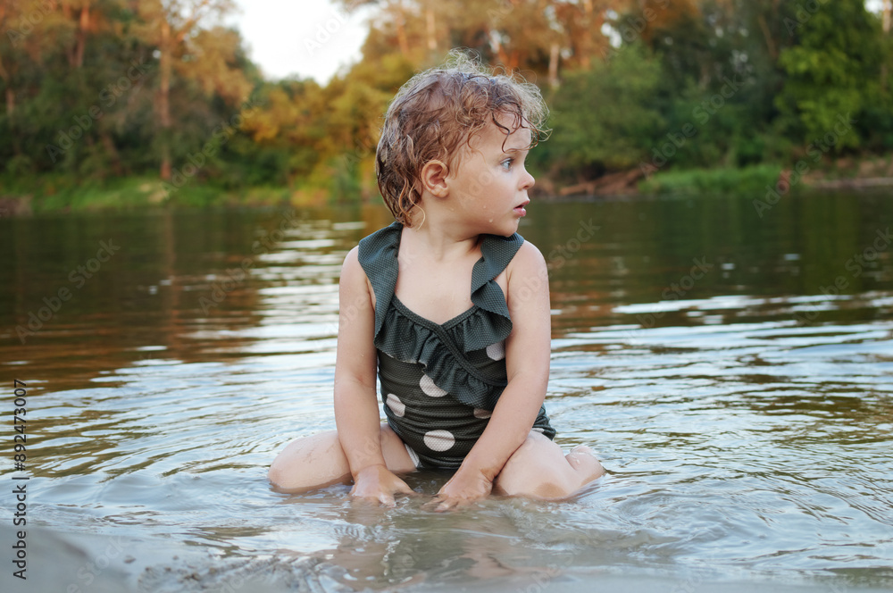 Light-haired curly Caucasian blonde little toddler girl dressed in dotted swimsuit sitting by the river splashing in the warm summer stream looking away at beautiful nature Light-haired curly Caucasian blonde little toddler girl dressed in dotted swimsuit sitting by the river splashing in the warm summer stream looking away at beautiful nature