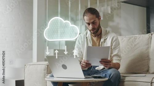 Modern Computer with Cloud Data Transfer Animation. Caucasian Man Working with Laptop and Documents at home Sitting on the Sofa.
