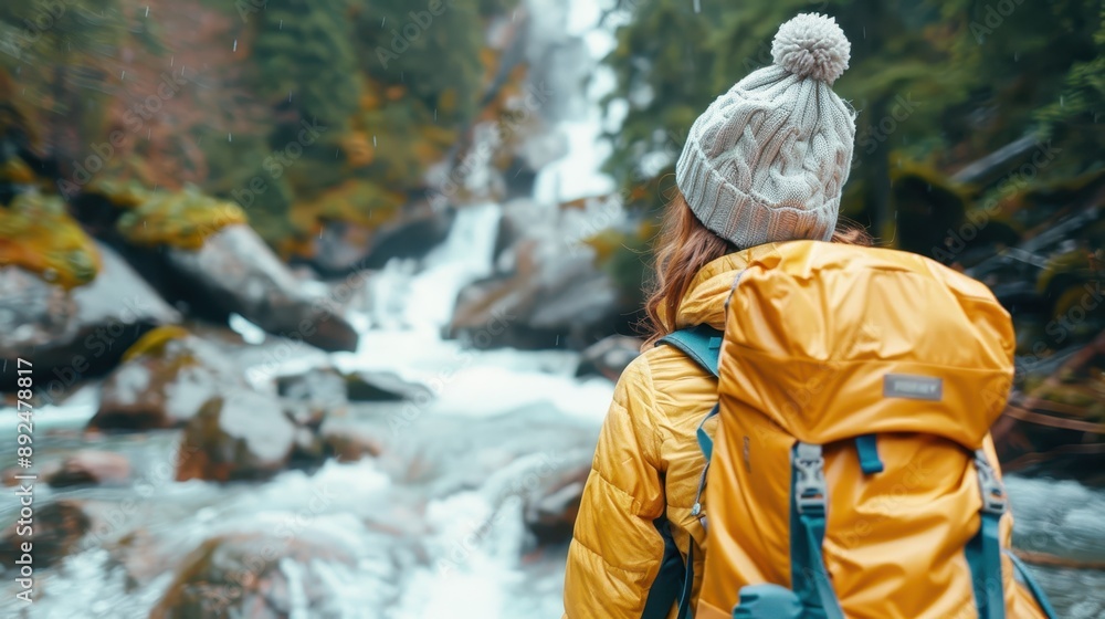 A hiker in a yellow jacket and gray cap with a backpack standing in front of a river waterfall, showcasing adventure and the majesty of nature in a forest setting. A hiker in a yellow jacket and gray cap with a backpack standing in front of a river waterfall, showcasing adventure and the majesty of nature in a forest setting.
