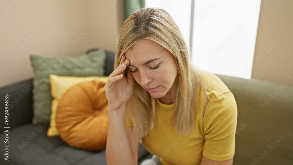 A concerned young blonde woman sitting indoors, touching her forehead in a gesture of worry or headache against a comfortable home background.