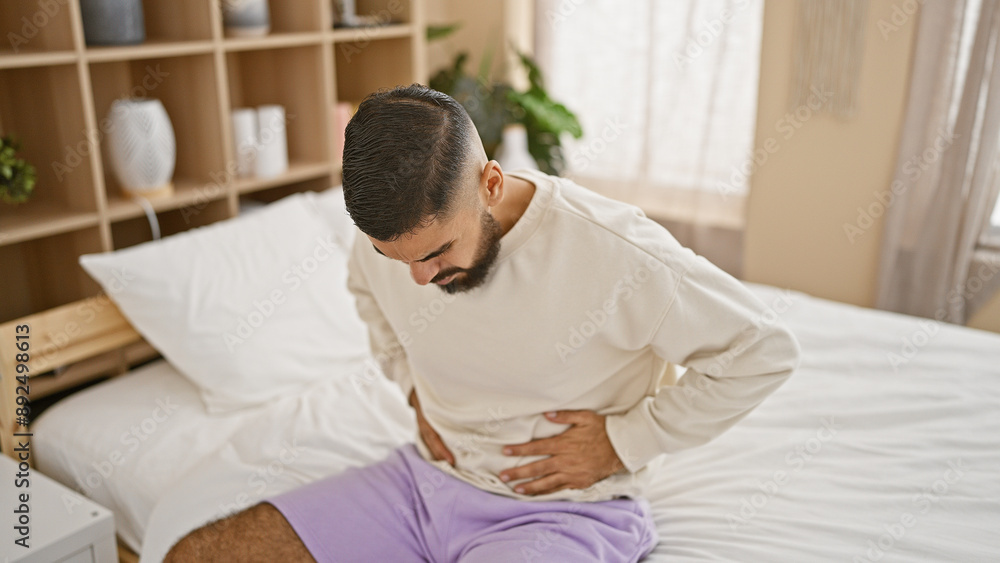 A bearded young man feeling stomach pain, sitting in his modern bedroom, reflects a moment of discomfort and worry. A bearded young man feeling stomach pain, sitting in his modern bedroom, reflects a moment of discomfort and worry.