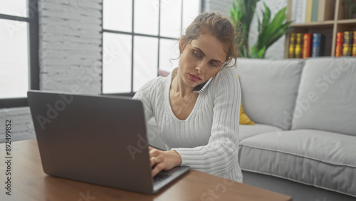 A young woman multitasks with a phone call and laptop work in a modern living room, exuding casual professional lifestyle vibes. photo