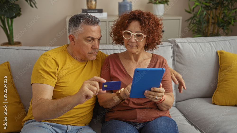Middle-aged couple sitting together on a couch at home using a tablet and credit card, showing love and teamwork in a modern indoor living room setting Middle-aged couple sitting together on a couch at home using a tablet and credit card, showing love and teamwork in a modern indoor living room setting