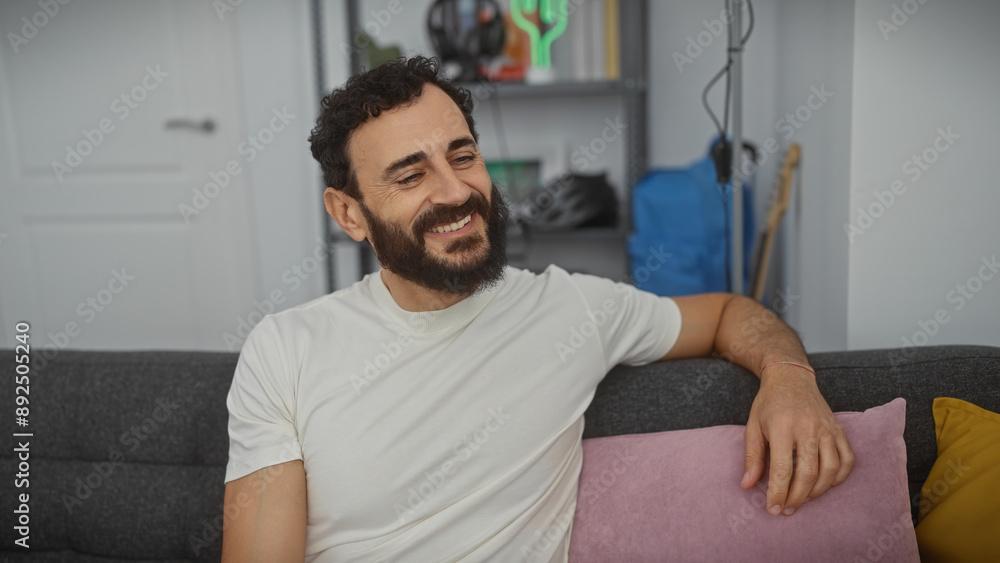 Smiling bearded man relaxing on a couch in a modern living room, exuding casual comfort and style. Smiling bearded man relaxing on a couch in a modern living room, exuding casual comfort and style.
