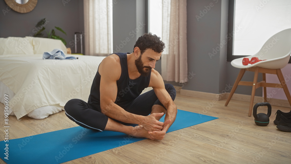 Bearded man stretching on yoga mat in bedroom with wooden floor and white bed Bearded man stretching on yoga mat in bedroom with wooden floor and white bed