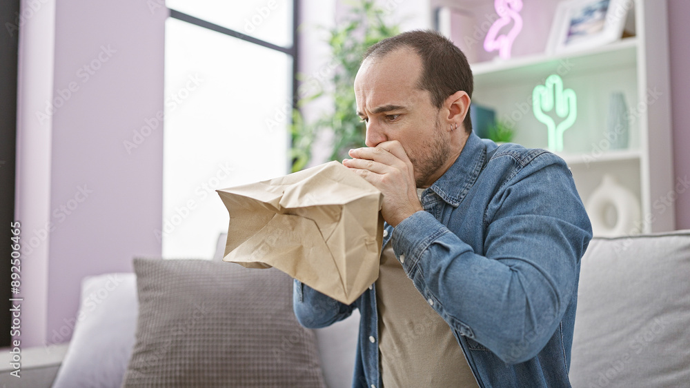 A bald man with a beard in a denim shirt breathes into a paper bag while sitting on a sofa indoors to calm an anxiety attack.