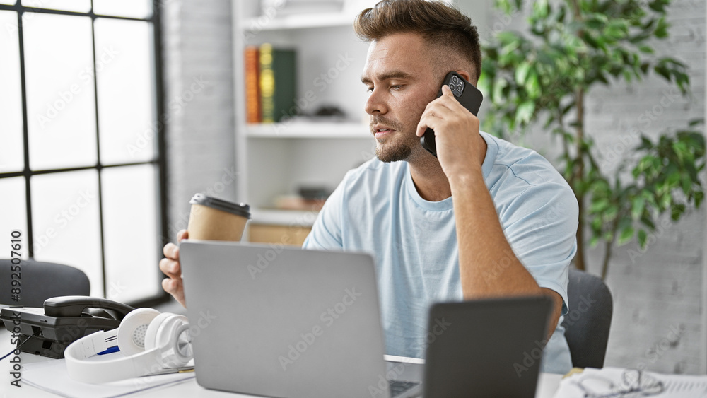 Handsome young man with beard in a modern office holding coffee and talking on smartphone while using laptop Handsome young man with beard in a modern office holding coffee and talking on smartphone while using laptop