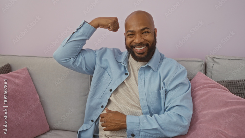 Handsome young black man with beard smiling and flexing arm while sitting in living room of home