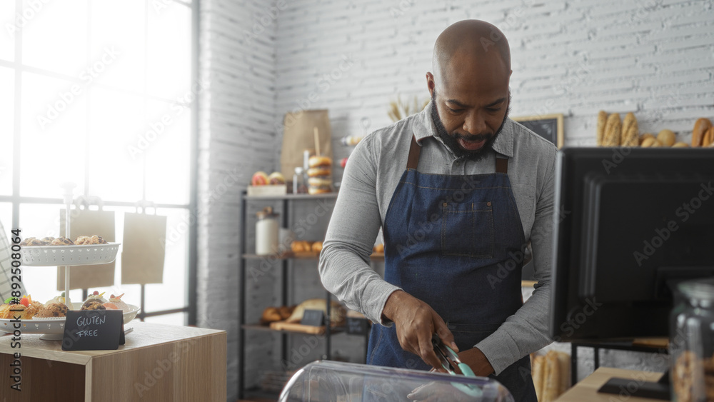 Handsome young african american man in a bakery shop wearing an apron while arranging desserts behind the counter in a bright, modern indoor setting Handsome young african american man in a bakery shop wearing an apron while arranging desserts behind the counter in a bright, modern indoor setting