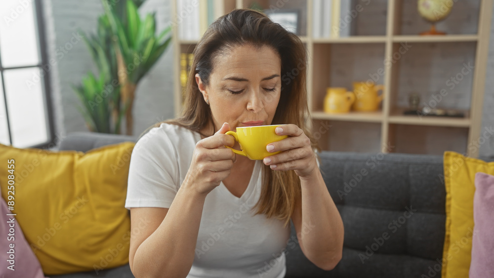 Hispanic woman enjoying coffee indoors on a cozy sofa with vibrant yellow accents in a modern living room setting. Hispanic woman enjoying coffee indoors on a cozy sofa with vibrant yellow accents in a modern living room setting.