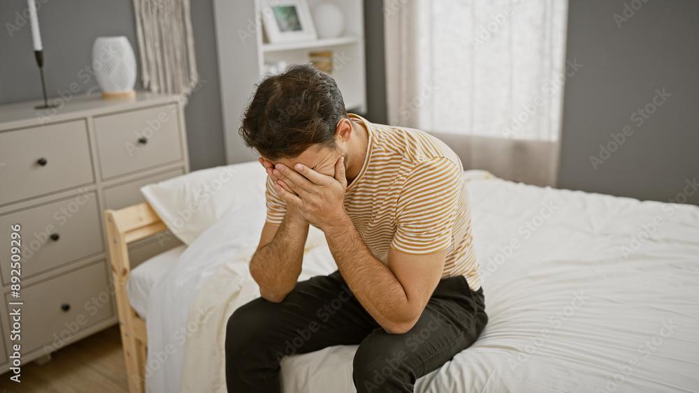 A distressed young hispanic man sits on a bed in a modern bedroom, covering his face with his hands. A distressed young hispanic man sits on a bed in a modern bedroom, covering his face with his hands.