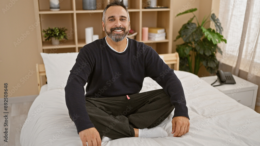 Fototapeta premium A mature hispanic man with grey beard smiling casually sitting on a white bed in a well-lit bedroom interior.