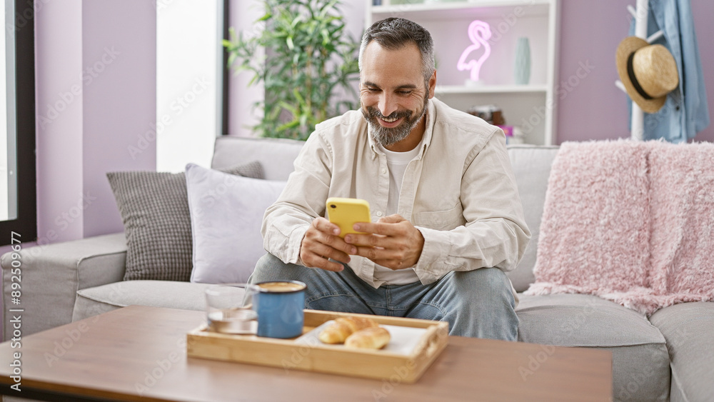 A smiling mature hispanic man with a beard and grey hair enjoys using a smartphone in a cozy living room setting. A smiling mature hispanic man with a beard and grey hair enjoys using a smartphone in a cozy living room setting.