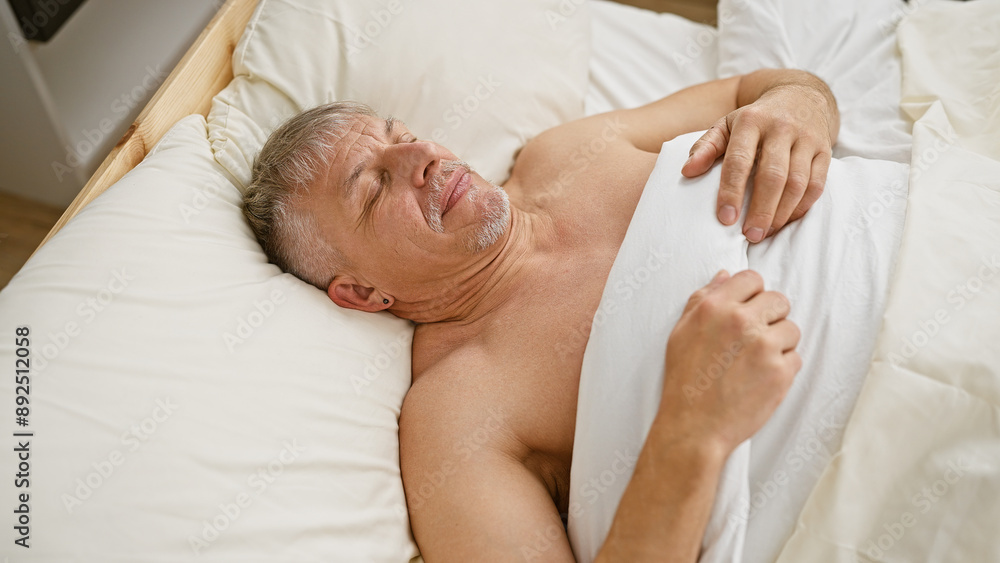A distressed middle-aged man lying in bed indoors grimacing as if experiencing pain, discomfort, or a bad dream. A distressed middle-aged man lying in bed indoors grimacing as if experiencing pain, discomfort, or a bad dream.
