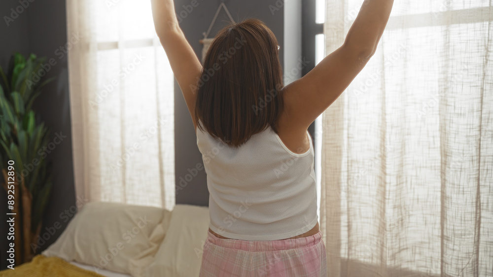 A middle-aged hispanic woman with short hair stretches her arms in the air while standing in her bedroom during the morning. A middle-aged hispanic woman with short hair stretches her arms in the air while standing in her bedroom during the morning.