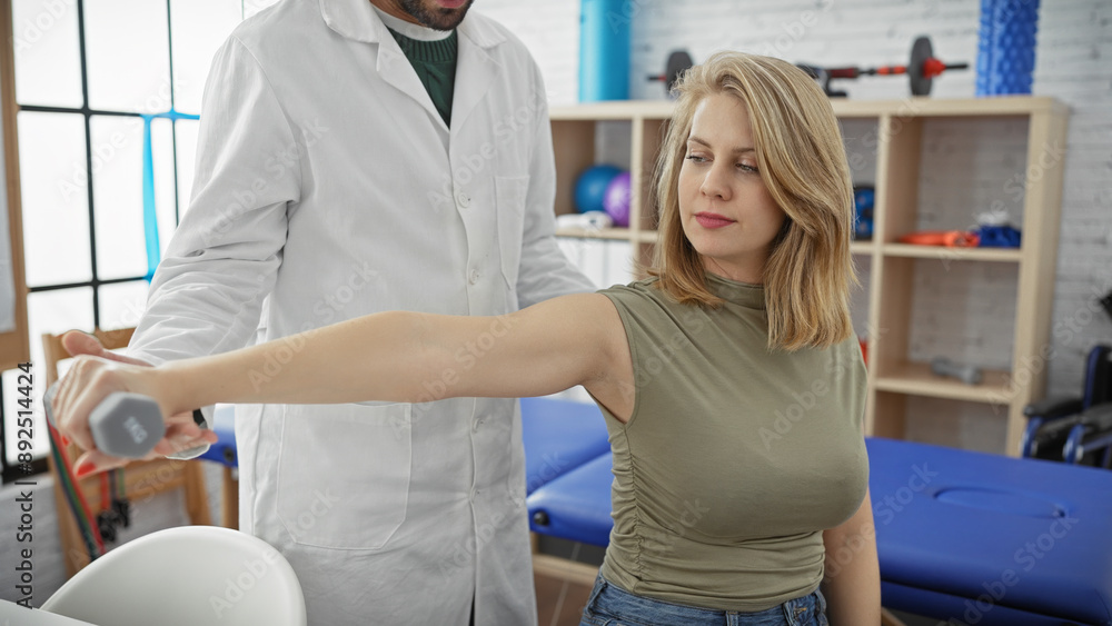 Obraz premium A woman exercises her arm with a dumbbell under a male physiotherapist's guidance in a well-equipped therapy room.
