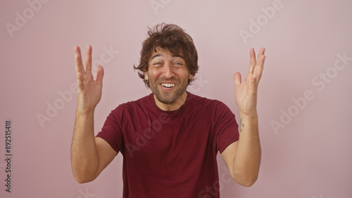 A cheerful young hispanic man with a beard, wearing a burgundy t-shirt, joyfully expresses excitement against a plain pink background.